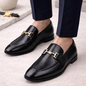May include: Black leather loafers with a gold-colored buckle detail. The shoes have a classic design with a rounded toe and a low heel. The loafers are shown with navy blue trousers.