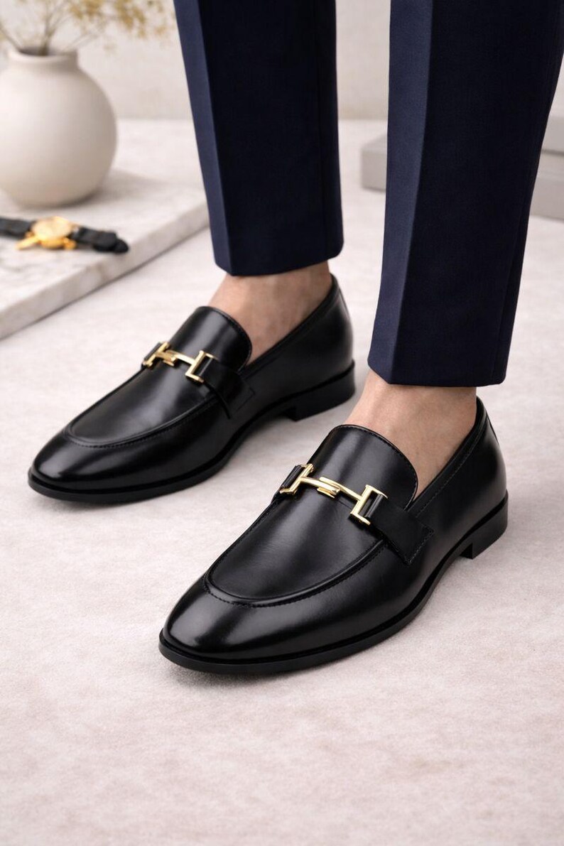 May include: Black leather loafers with a gold-colored buckle detail. The shoes have a classic design with a rounded toe and a low heel. The loafers are shown with navy blue trousers.
