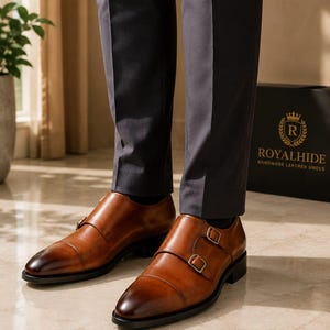 May include: A pair of tan leather double monk strap shoes with a burnished toe finish, silver-toned metal buckles, and a black sole. The shoes are worn with dark grey trousers, and a black box with the text ROYALHIDE HANDMADE LEATHER SHOES is visible.