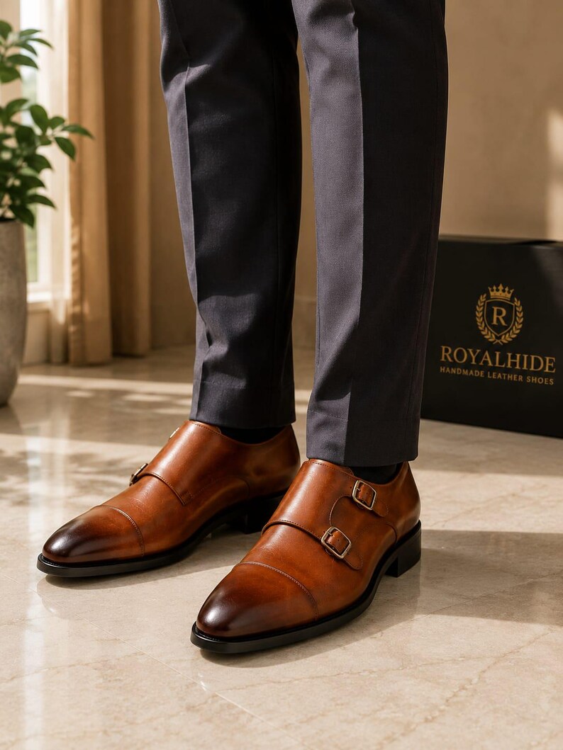 May include: A pair of tan leather double monk strap shoes with a burnished toe finish, silver-toned metal buckles, and a black sole. The shoes are worn with dark grey trousers, and a black box with the text ROYALHIDE HANDMADE LEATHER SHOES is visible.