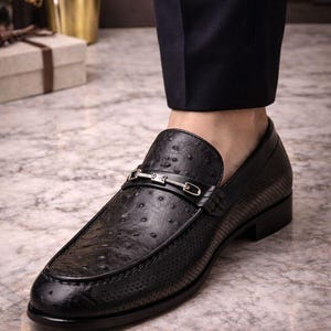 Pu&ograve; includere: Mocassino in pelle nera con motivo a trama di struzzo e fibbia color argento. La scarpa presenta un design traforato sulla tomaia e un tacco basso. La scarpa &egrave; abbinata a pantaloni blu navy scuro.