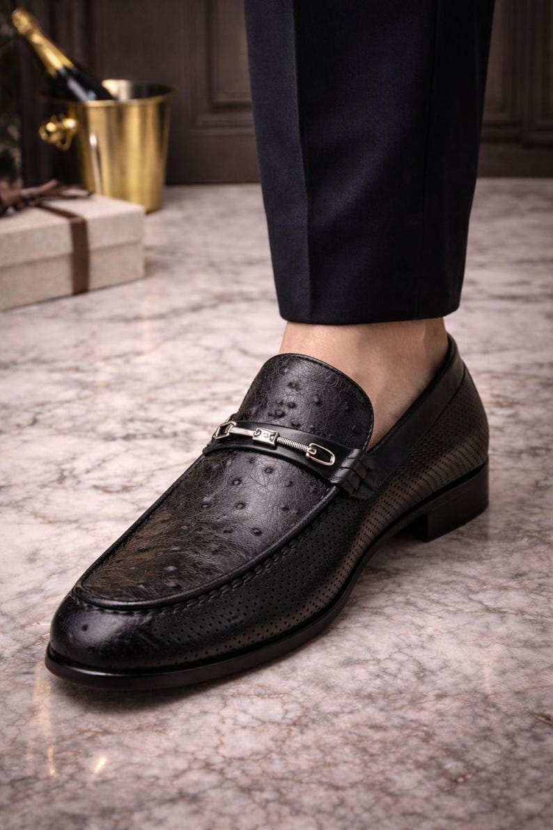 Pu&ograve; includere: Mocassino in pelle nera con motivo a trama di struzzo e fibbia color argento. La scarpa presenta un design traforato sulla tomaia e un tacco basso. La scarpa &egrave; abbinata a pantaloni blu navy scuro.