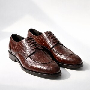 K&ouml;nnte beinhalten: Ein Paar braune Lederschuhe mit Krokodiltextur. Die Schuhe haben ein klassisches Schn&uuml;rdesign und ein poliertes Finish, geeignet f&uuml;r formelle Anl&auml;sse. Die Schuhe werden auf wei&szlig;em Hintergrund pr&auml;sentiert.