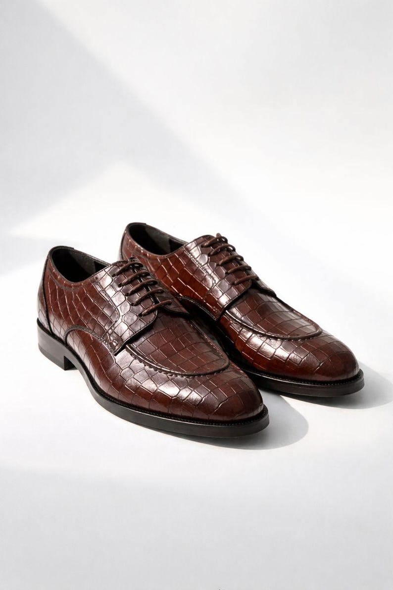 K&ouml;nnte beinhalten: Ein Paar braune Lederschuhe mit Krokodiltextur. Die Schuhe haben ein klassisches Schn&uuml;rdesign und ein poliertes Finish, geeignet f&uuml;r formelle Anl&auml;sse. Die Schuhe werden auf wei&szlig;em Hintergrund pr&auml;sentiert.
