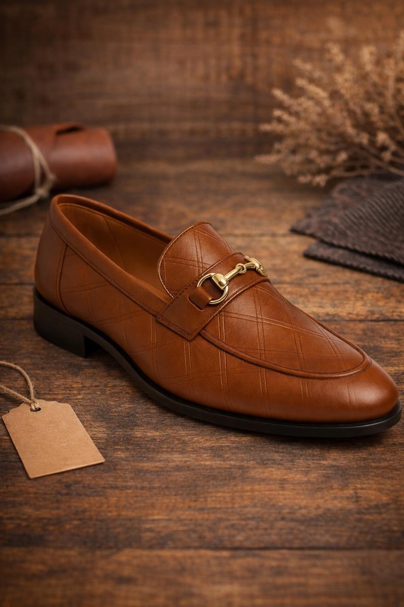 May include: A brown leather loafer with a gold-colored buckle. The shoe has a diamond pattern on the upper and a black sole. The shoe is displayed on a wooden surface.