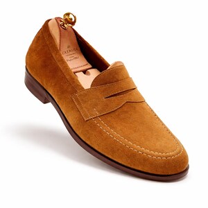 May include: A single brown suede loafer with a penny strap design. The shoe features a stitched sole and a wooden shoe tree inside. The loafer is a warm tan color.