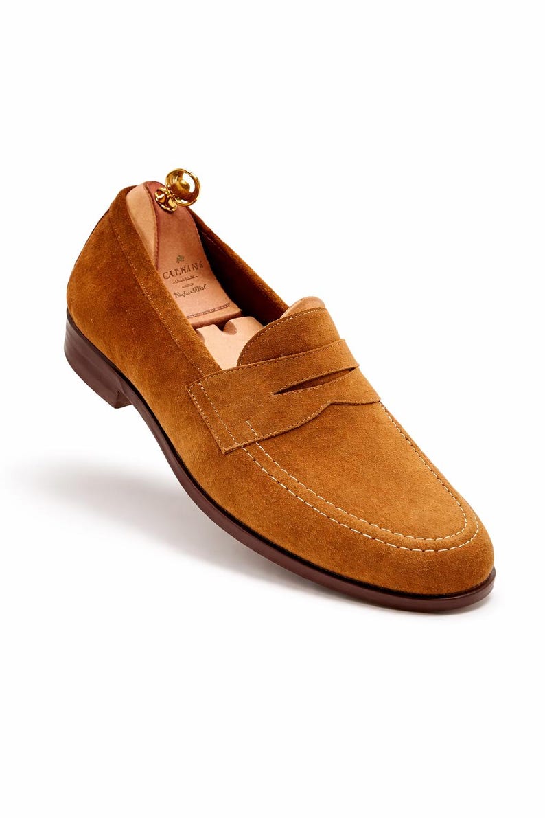 May include: A single brown suede loafer with a penny strap design. The shoe features a stitched sole and a wooden shoe tree inside. The loafer is a warm tan color.