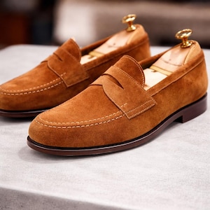 May include: A pair of brown suede penny loafers with a classic design. The shoes feature a rounded toe, a strap across the vamp, and a brown leather sole. The shoes are displayed with wooden shoe trees.