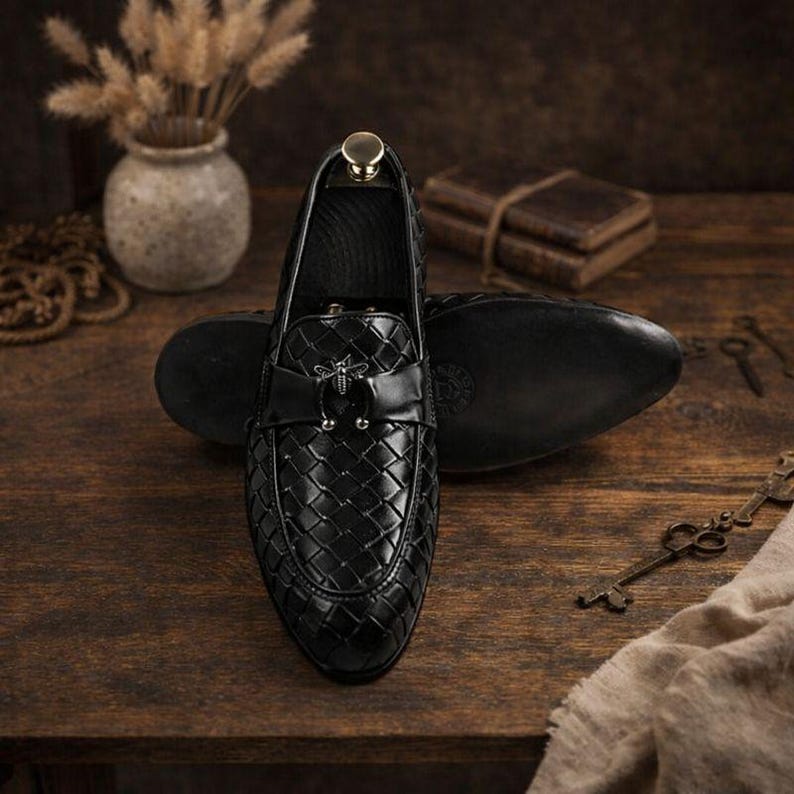 May include: Black leather loafers with a woven pattern and a decorative bee emblem. The shoes are displayed on a wooden surface with a blurred background, showcasing a classic and elegant design. The loafers are a size 10.