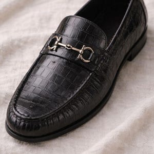 May include: Black leather loafer with a crocodile texture. The shoe features a silver-colored metal strap across the top. The shoe is on a white fabric surface. A classic style of footwear.