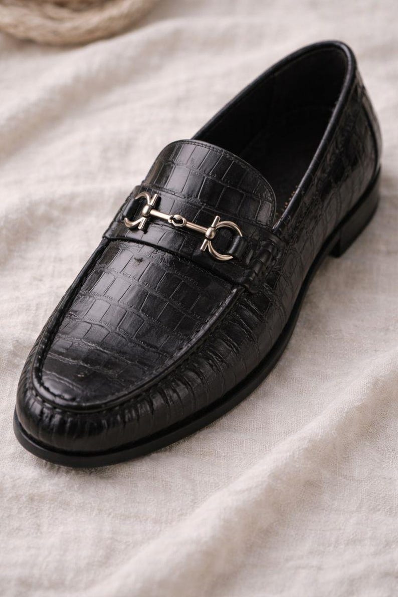 May include: Black leather loafer with a crocodile texture. The shoe features a silver-colored metal strap across the top. The shoe is on a white fabric surface. A classic style of footwear.