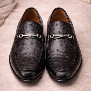 Pu&ograve; includere: Un paio di mocassini in pelle nera con un motivo strutturato e puntinato. Ogni scarpa presenta un dettaglio con fibbia color argento sulla parte superiore. Le scarpe sono esposte su una superficie dai toni neutri.