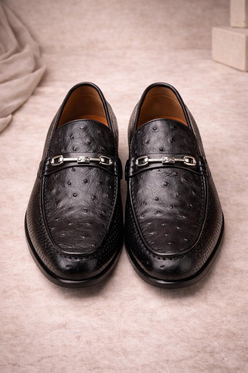 Pu&ograve; includere: Un paio di mocassini in pelle nera con un motivo strutturato e puntinato. Ogni scarpa presenta un dettaglio con fibbia color argento sulla parte superiore. Le scarpe sono esposte su una superficie dai toni neutri.