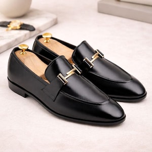 May include: A pair of black leather loafers with gold-tone metal accents. The shoes have a classic design with a rounded toe and a decorative strap across the top. The loafers are displayed on a light-colored surface.