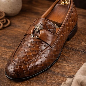 May include: Brown leather loafer with a woven pattern. The shoe features a decorative strap with a metal accent. The shoe is on a wooden shoe form. The shoe is on a wooden surface.