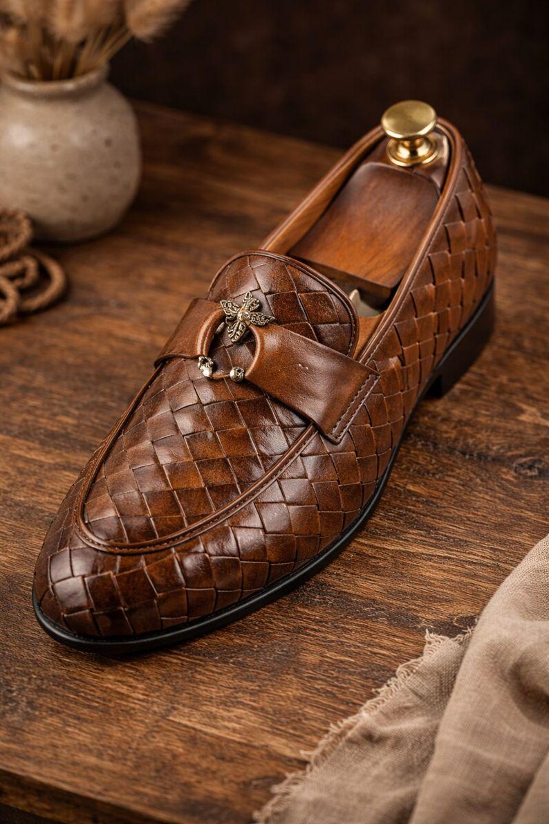 May include: Brown leather loafer with a woven pattern. The shoe features a decorative strap with a metal accent. The shoe is on a wooden shoe form. The shoe is on a wooden surface.
