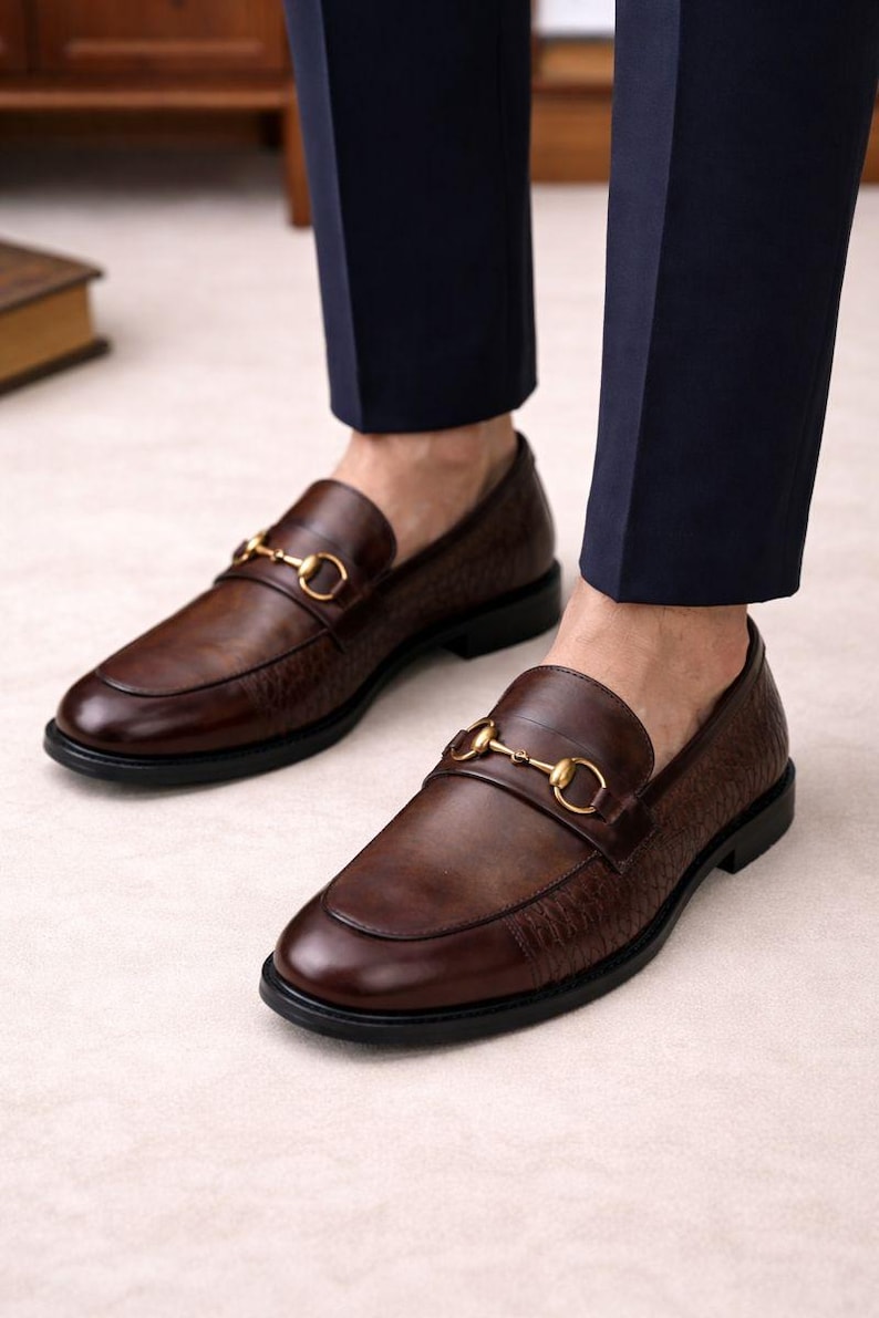 Puede incluir: Mocasines de cuero marr&oacute;n con detalles dorados de adorno de caballo. Los zapatos tienen una parte superior texturizada y una suela negra. La imagen muestra los zapatos desde abajo, destacando su dise&ntilde;o.
