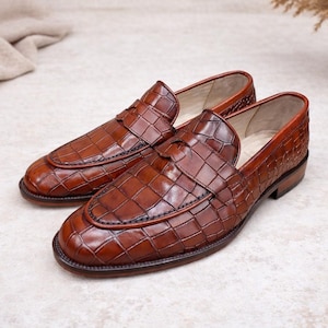 May include: A pair of brown leather loafers with a textured, crocodile-like pattern. The shoes have a classic design with a rounded toe and a low heel. The leather has a glossy finish, and the shoes are displayed on a light-colored surface.