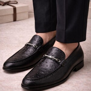 Pu&ograve; includere: Mocassini in pelle nera con un design strutturato e traforato. Ogni scarpa presenta una cinghia in metallo color argento. Indossati con pantaloni neri, suggeriscono un ambiente formale o professionale.