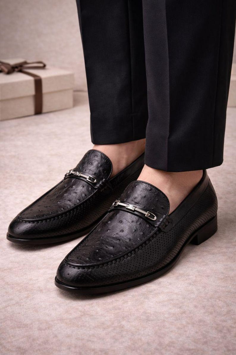 Pu&ograve; includere: Mocassini in pelle nera con un design strutturato e traforato. Ogni scarpa presenta una cinghia in metallo color argento. Indossati con pantaloni neri, suggeriscono un ambiente formale o professionale.