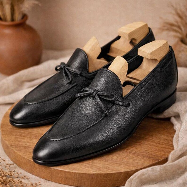 May include: A pair of black leather loafers with a textured finish, featuring a bow detail on the vamp. The shoes are displayed with wooden shoe trees on a wooden surface, showcasing a classic design.