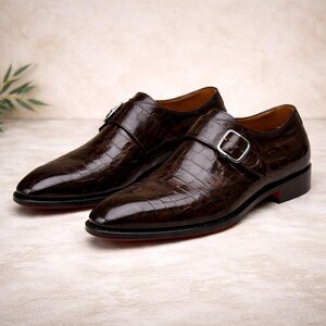 May include: A pair of brown leather dress shoes with a crocodile texture. The shoes feature a silver buckle closure and a red sole. The shoes are displayed on a neutral background.