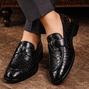 May include: Black leather loafers with a textured, quilted pattern. Each shoe features a decorative silver-tone buckle. The shoes are worn with dark gray dress pants, suggesting a formal or business attire.