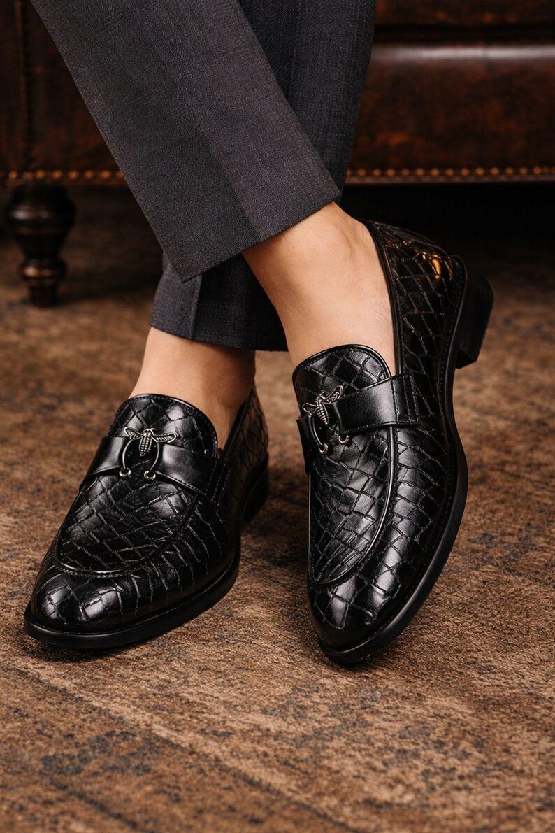 May include: Black leather loafers with a textured, quilted pattern. Each shoe features a decorative silver-tone buckle. The shoes are worn with dark gray dress pants, suggesting a formal or business attire.