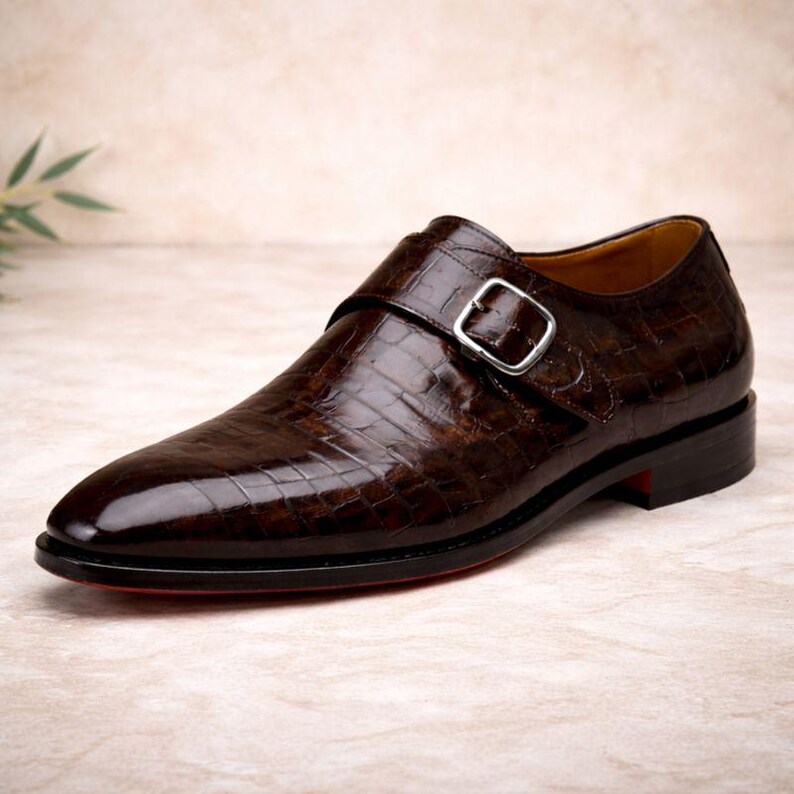 May include: A brown leather monk strap shoe with a textured, crocodile-like pattern. The shoe features a silver buckle, a pointed toe, and a red sole. The shoe is a size 10.