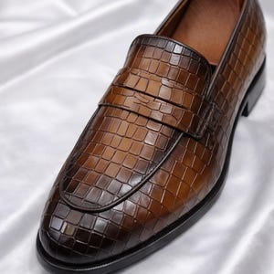 K&ouml;nnte beinhalten: Brauner Leder-Loafer mit einem strukturierten, krokodilartigen Muster. Der Schuh hat ein klassisches Design mit runder Spitze und einem Riemen &uuml;ber dem Spann. Der Schuh befindet sich auf einer wei&szlig;en Satin-Oberfl&auml;che.