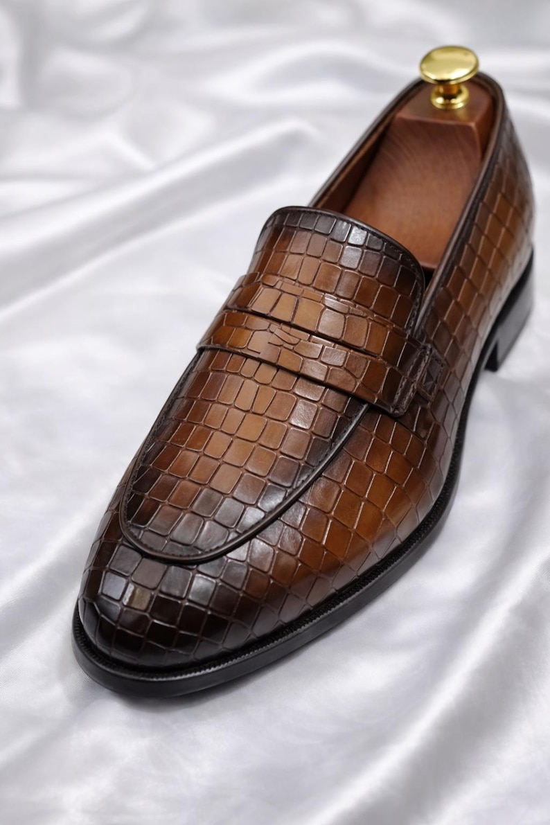 K&ouml;nnte beinhalten: Brauner Leder-Loafer mit einem strukturierten, krokodilartigen Muster. Der Schuh hat ein klassisches Design mit runder Spitze und einem Riemen &uuml;ber dem Spann. Der Schuh befindet sich auf einer wei&szlig;en Satin-Oberfl&auml;che.