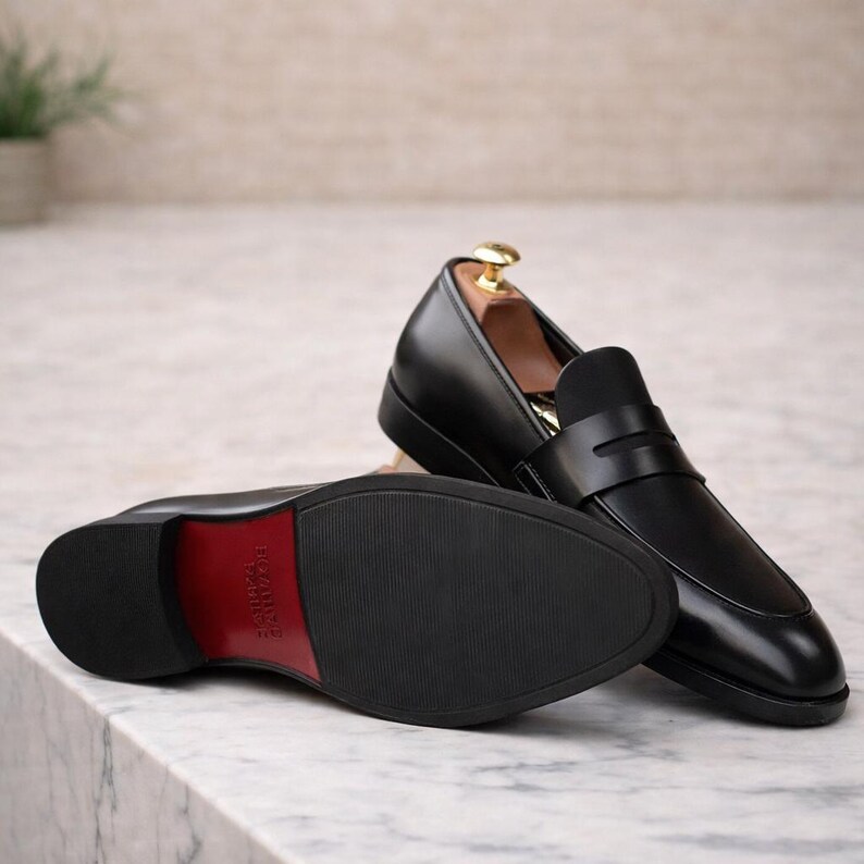 May include: Black leather penny loafers with a red sole accent. One shoe is upright, showcasing the design, while the other lies on its side, revealing the sole's texture and color. The shoes are displayed on a marble surface.