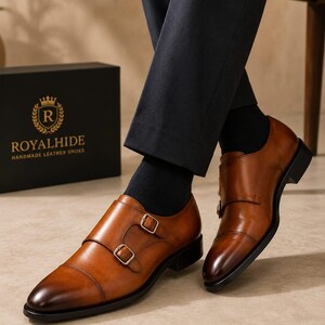 May include: A pair of tan leather double monk strap shoes with a burnished toe, featuring two gold-toned metal buckles on each shoe, a black sole, and a black heel. The shoes are worn with dark trousers and black socks, near a black box with the text ROYALHIDE.