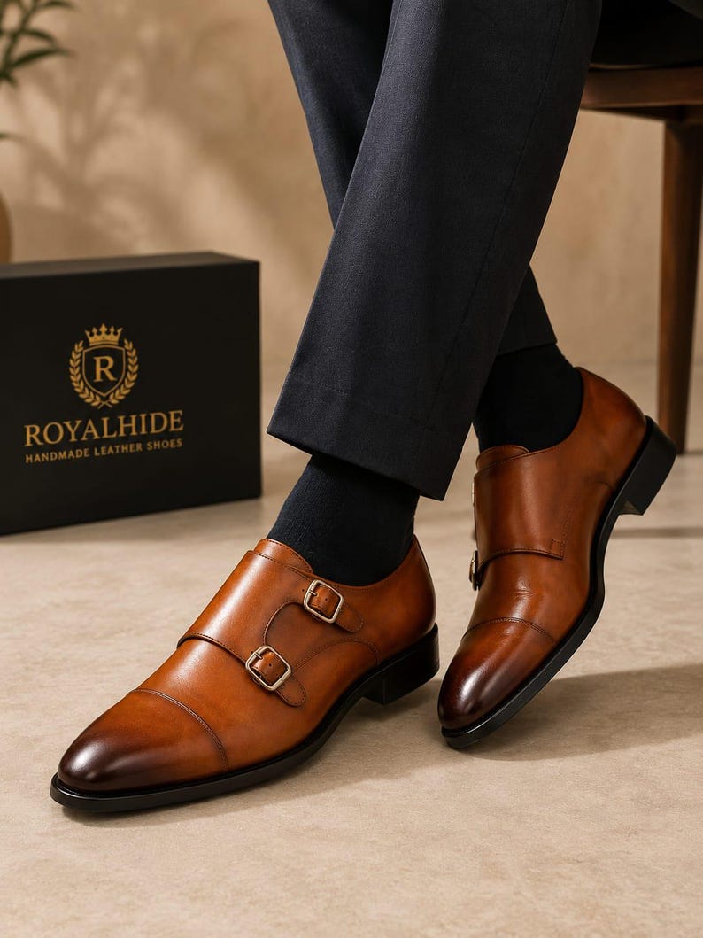 May include: A pair of tan leather double monk strap shoes with a burnished toe, featuring two gold-toned metal buckles on each shoe, a black sole, and a black heel. The shoes are worn with dark trousers and black socks, near a black box with the text ROYALHIDE.