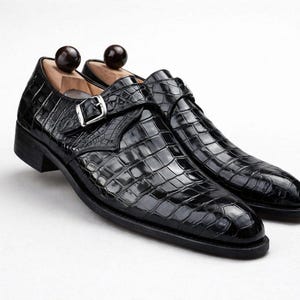 Op de afbeelding: Een paar zwarte leren schoenen met een krokodillenleertextuur. De schoenen hebben een gespsluiting en worden gepresenteerd met houten schoenspanners. Op een witte achtergrond.