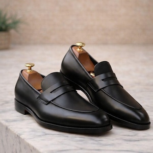 May include: A pair of black leather penny loafers with a classic design. The shoes have a smooth, polished finish and a strap across the vamp. Wooden shoe trees are inside the shoes. The loafers are displayed on a marble surface.