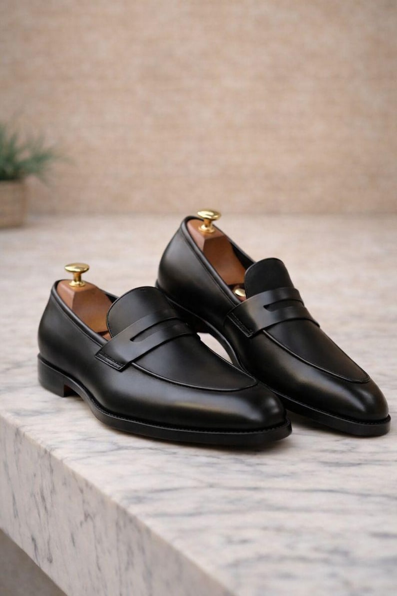 May include: A pair of black leather penny loafers with a classic design. The shoes have a smooth, polished finish and a strap across the vamp. Wooden shoe trees are inside the shoes. The loafers are displayed on a marble surface.