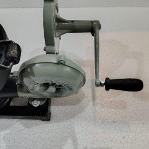 Puede incluir: Soplador de forja de hierro fundido gris claro con manivela. El soplador tiene una carcasa redonda con el texto "AZAT FACTORY" y un mango negro. Est&aacute; montado sobre una base negra.
