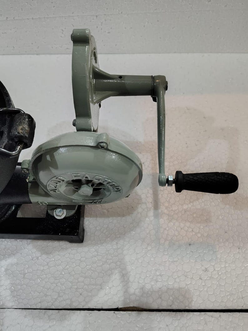 Puede incluir: Soplador de forja de hierro fundido gris claro con manivela. El soplador tiene una carcasa redonda con el texto "AZAT FACTORY" y un mango negro. Est&aacute; montado sobre una base negra.