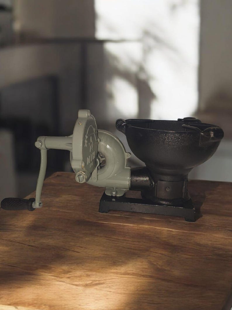 Puede incluir: Un molinillo de caf&eacute; antiguo con manivela, con cuerpo gris y tolva negra. El molinillo se encuentra sobre una superficie de madera, mostrando su dise&ntilde;o cl&aacute;sico y funcionalidad. El mango es negro y el molinillo tiene un dise&ntilde;o de estrella en el lateral.