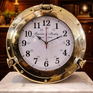 Könnte beinhalten: Eine messingfarbene Uhr mit weißem Zifferblatt und schwarzen Ziffern. Die Uhr hat eine runde Form mit einem aufklappbaren Deckel und ist mit "Franklin & Murphy EST 1868" gekennzeichnet. Die Uhr steht auf einer Marmoroberfläche.