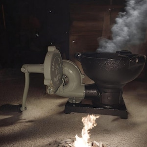 Puede incluir: Un fuelle de manivela gris verdoso vintage y un brasero de hierro fundido negro con un peque&ntilde;o fuego ardiendo. El humo se eleva del pozo. El fuelle tiene un asa y un dise&ntilde;o decorativo. El brasero est&aacute; sobre una base negra.