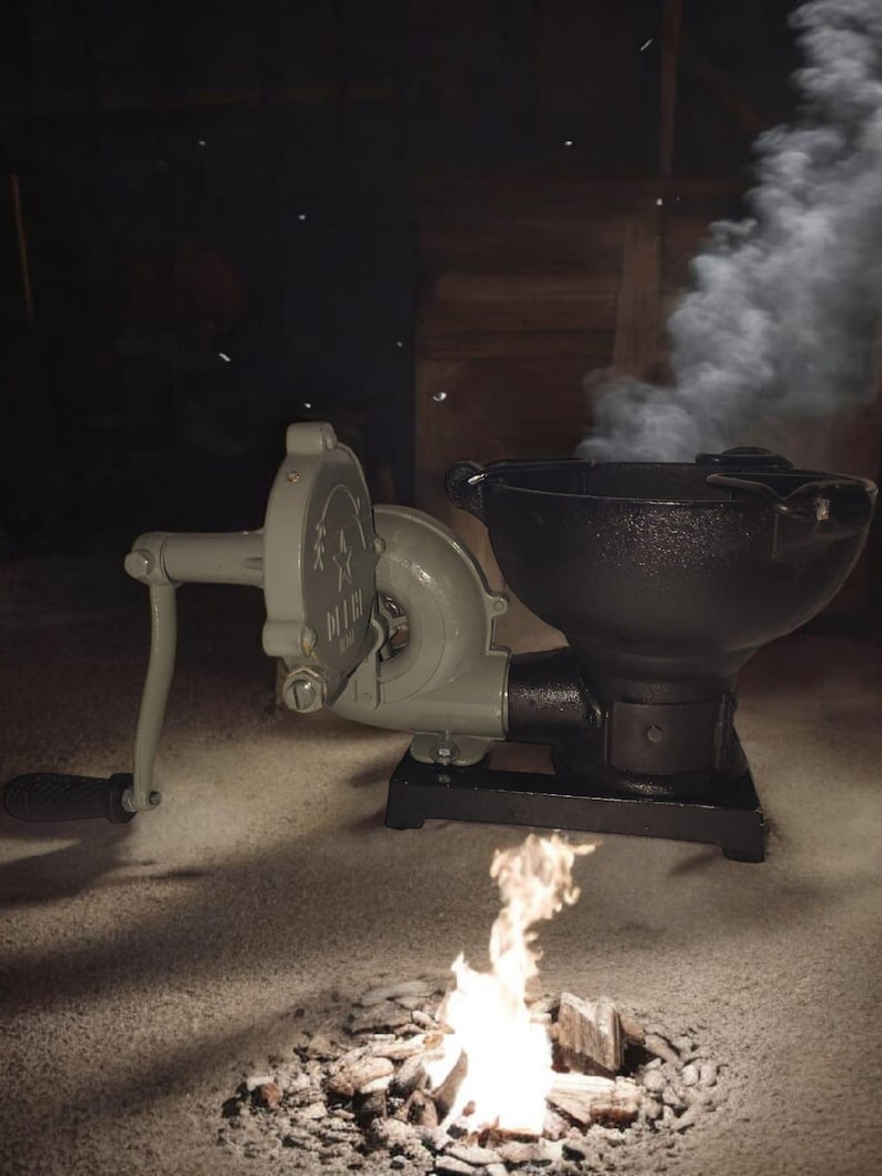 Puede incluir: Un fuelle de manivela gris verdoso vintage y un brasero de hierro fundido negro con un peque&ntilde;o fuego ardiendo. El humo se eleva del pozo. El fuelle tiene un asa y un dise&ntilde;o decorativo. El brasero est&aacute; sobre una base negra.