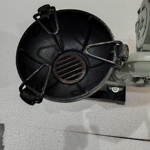 Puede incluir: Un objeto met&aacute;lico negro y redondo con un asa y una rejilla. El objeto est&aacute; fijado a una base met&aacute;lica gris claro. Parece ser parte de una m&aacute;quina o aparato m&aacute;s grande.