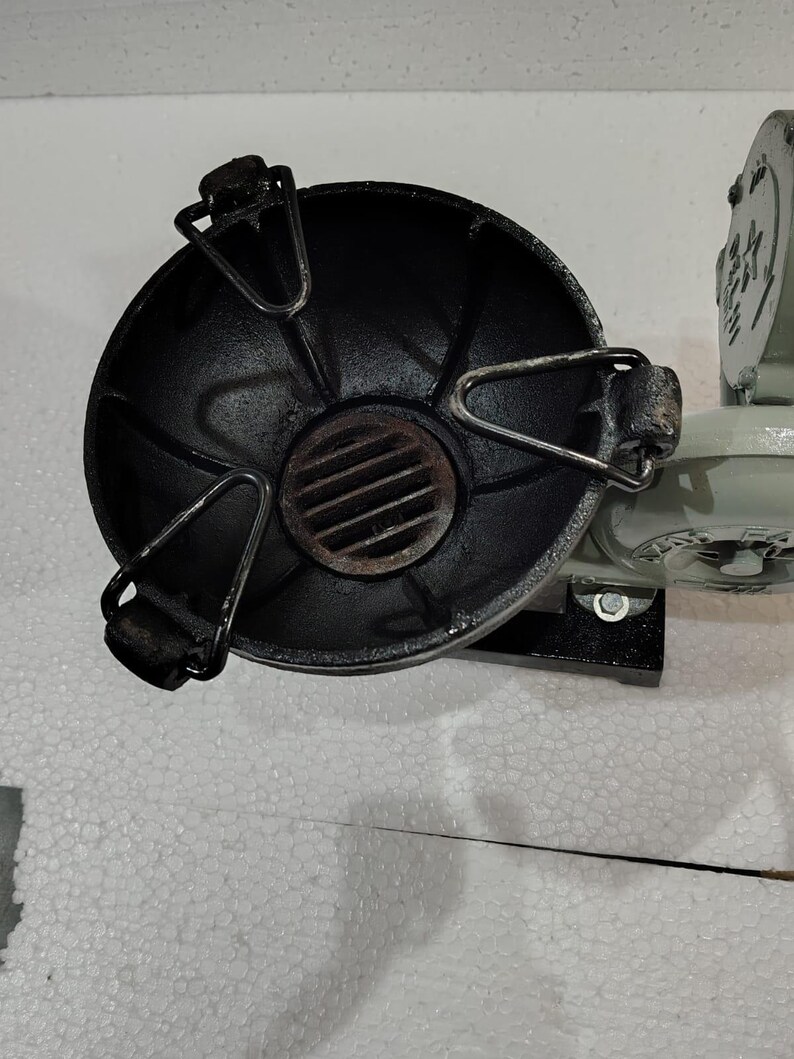 Puede incluir: Un objeto met&aacute;lico negro y redondo con un asa y una rejilla. El objeto est&aacute; fijado a una base met&aacute;lica gris claro. Parece ser parte de una m&aacute;quina o aparato m&aacute;s grande.