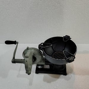 Puede incluir: Un brasero de metal color carb&oacute;n con un soplador de manivela. El brasero tiene forma de cuenco redondo con una rejilla en el interior. El soplador es gris con un mango negro. El brasero y el soplador est&aacute;n conectados por un marco de metal.