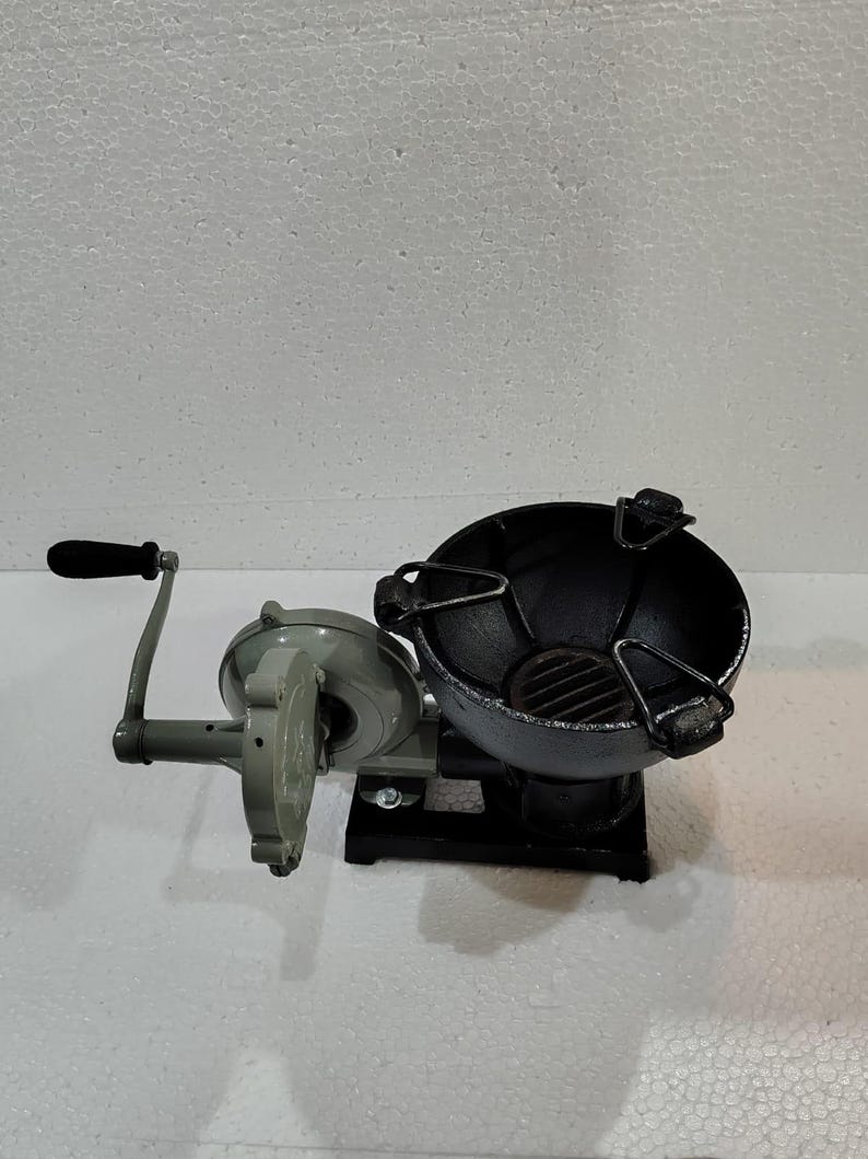 Puede incluir: Un brasero de metal color carb&oacute;n con un soplador de manivela. El brasero tiene forma de cuenco redondo con una rejilla en el interior. El soplador es gris con un mango negro. El brasero y el soplador est&aacute;n conectados por un marco de metal.