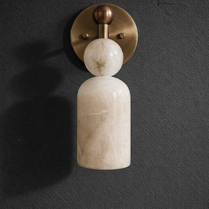 May include: A wall sconce featuring a circular antique brass backplate with a central stem. Attached to the stem is a spherical white stone orb positioned above a larger, cylindrical white stone body with subtle beige veining throughout.