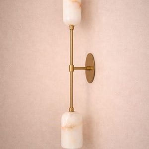 Peut inclure: Applique murale moderne avec abat-jour cylindriques en albâtre blanc cassé et finition laiton. Le luminaire a un design vertical avec deux abat-jours en albâtre et une plaque murale circulaire. L'albâtre a une teinte rosée subtile et chaleureuse.