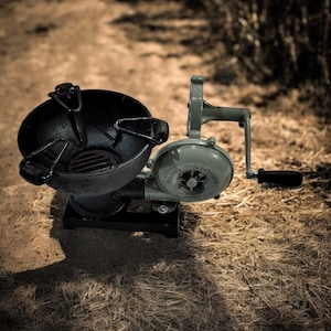 Puede incluir: Un soplador de forja vintage, accionado a mano, pintado en verde oliva y negro. El soplador presenta una carcasa de ventilador circular con el texto "Hazard Factory" y un mango para su funcionamiento. La forja tiene un hogar negro en forma de cuenco.